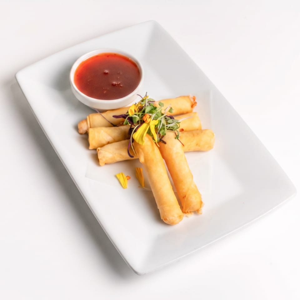 Golden-brown spring rolls with microgreens and red dipping sauce on a white rectangular plate.