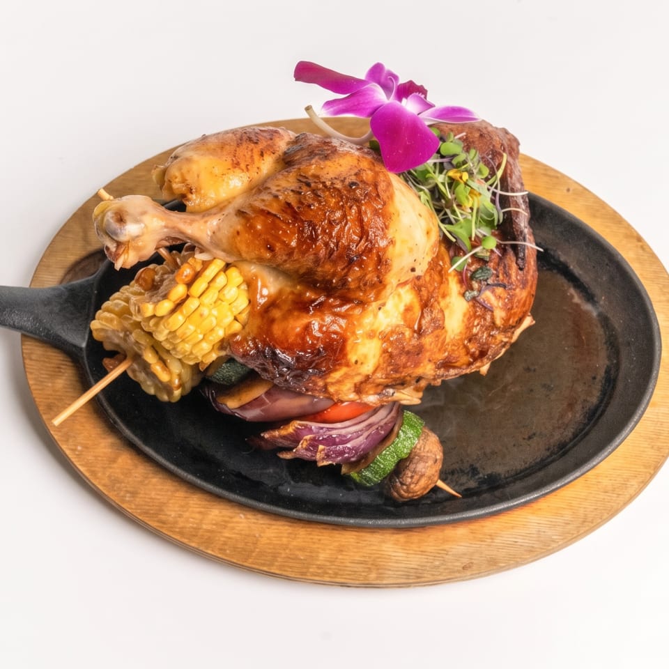 Whole roasted chicken on a sizzling plate with corn, vegetable skewer, microgreens, and an orchid.