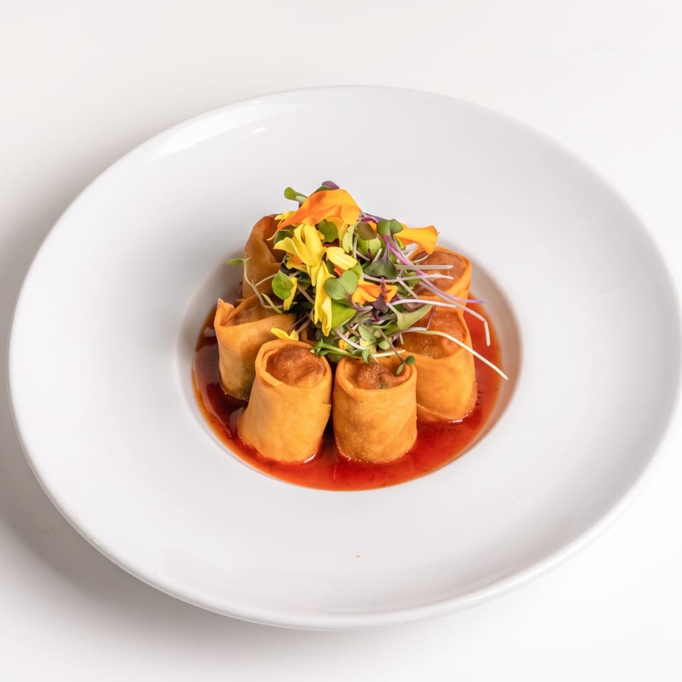 Appetizer of golden spring rolls in red sauce, topped with edible flowers and microgreens.